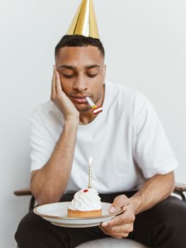 A man in a party hat looking at a cupcake with a lit candle in a contemplative mood.