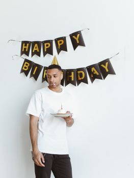 African American man holding birthday cake in front of festive banner, wearing party hat indoors.