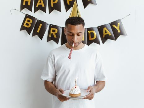 Man in party hat holding a cupcake with candle alone indoors. Birthday theme.