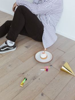 A man sitting alone with a birthday cupcake and party items on a wooden floor, marking a solitary celebration.