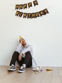 A man sits alone beneath a 'Happy Birthday' banner, wearing a party hat.