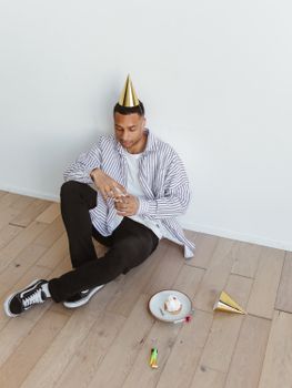 An adult man sitting alone with a party hat and a slice of cake, conveying a sense of loneliness during a celebration.