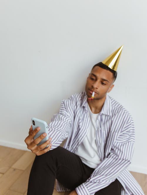 Man in White and Black Striped Dress Shirt Sitting on the Floor Looking at His Cellphone
