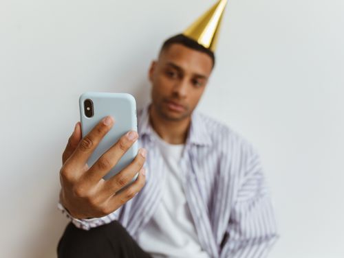 Free A man wearing a party hat takes a selfie with his smartphone indoors against a plain white background. Stock Photo