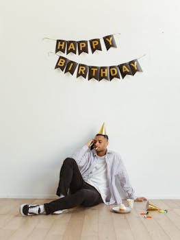 Man in a birthday hat sitting alone by a Happy Birthday banner, talking on the phone indoors.