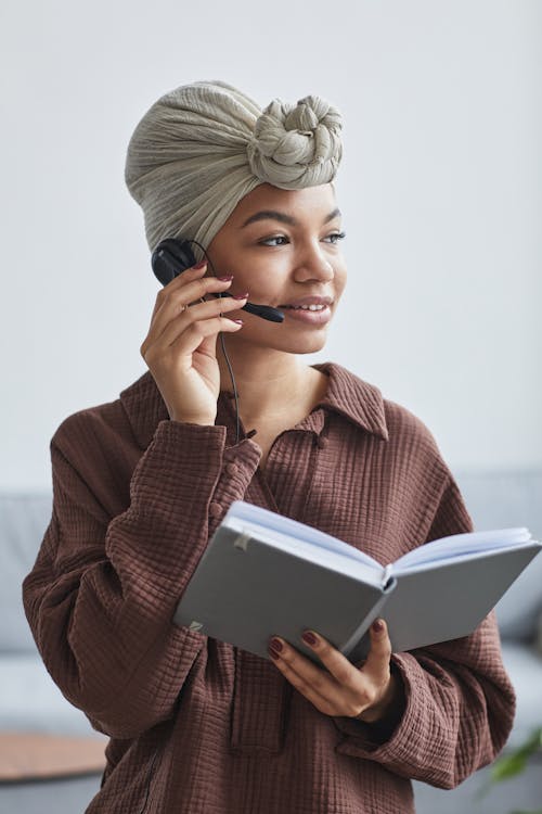 Free Positive African American female call agent in headset wearing turban standing in light room with notepad in light room at home Stock Photo