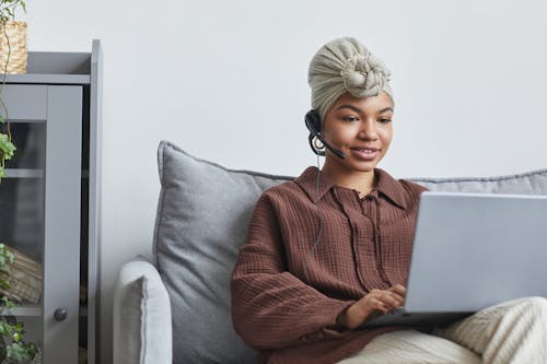 Free Focused African American female call agent with handsfree headset typing on netbook while sitting on couch near cupboard in light room Stock Photo