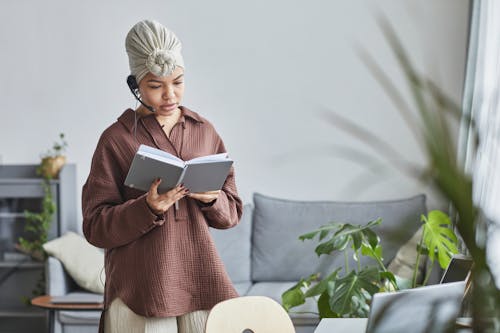 Free Focused African American female call agent with microphone in turban reading notes in notepad during work in room with blurred plants Stock Photo