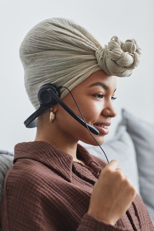 Free Side view of positive African American call agent in turban with microphone sitting on couch in light room at home Stock Photo