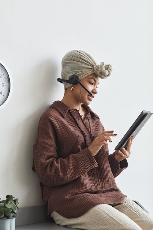 Free Focused African American female call agent with microphone browsing tablet while sitting at white wall during work in light room Stock Photo