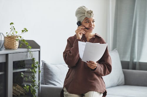 Free African American female call agent with headset and documents sitting on couch during conversation in light room with cupboard and plant Stock Photo