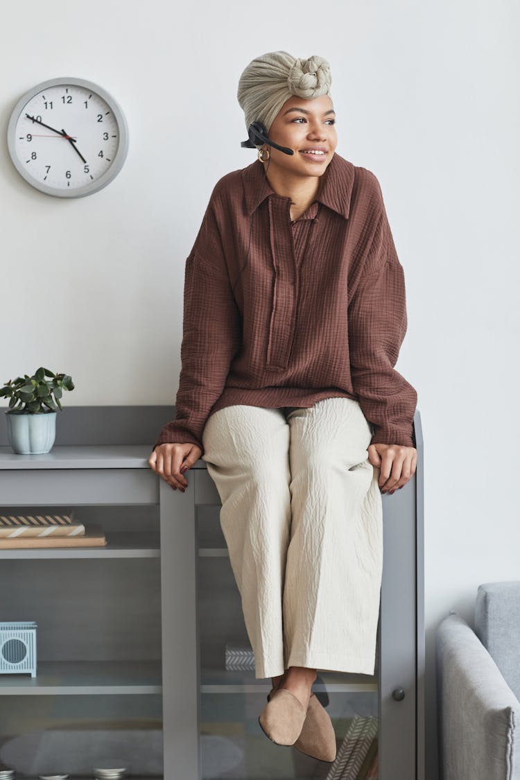 Positive Black Woman In Headset Sitting On Cupboard