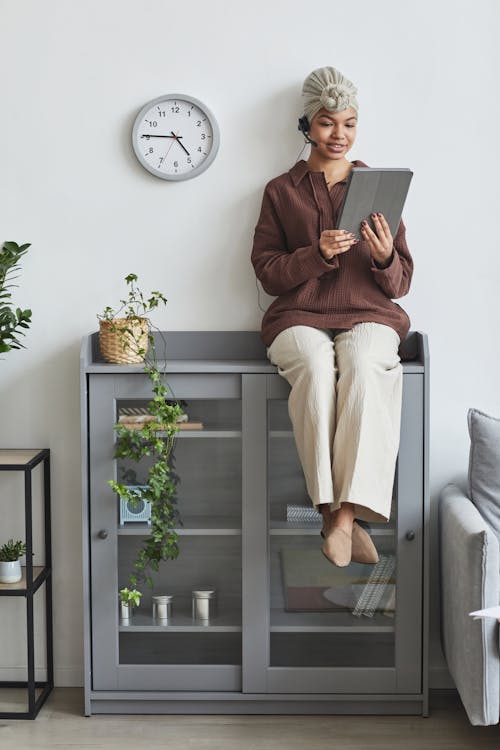 Black woman with headset and tablet sitting on cupboard