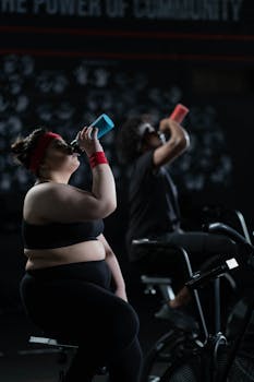 Two individuals on exercise bikes in a gym, staying hydrated, embracing body positivity.