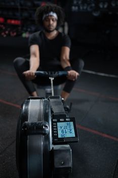 A focused man exercising on a rowing machine in a dimly lit gym setting, emphasizing physical fitness.