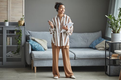 Smiling Woman in Headphones and with Notebook Standing by Couch