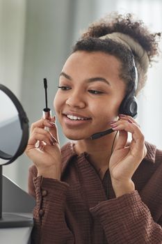 Smiling young woman wearing a headset and applying makeup in front of a mirror.