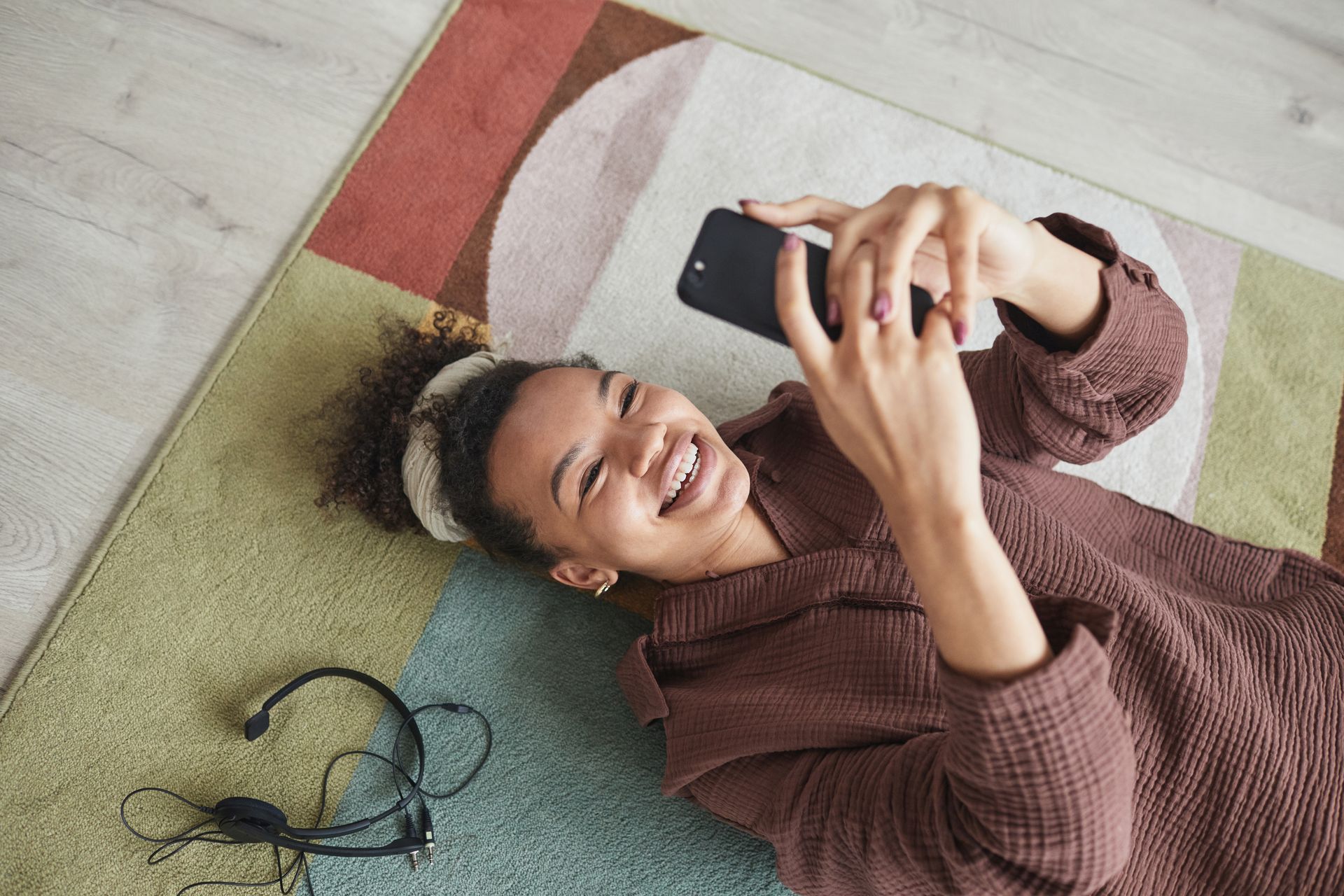 Smiling woman lying on a rug, enjoying relaxing time and texting on her smartphone indoors.