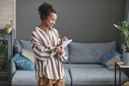 Woman wearing a headset, smiling and writing notes while working from home, exemplifying modern remote work.