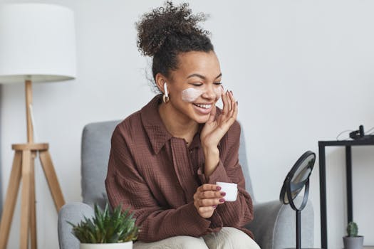 Smiling woman with curly hair applying face cream in a cozy living room setting.