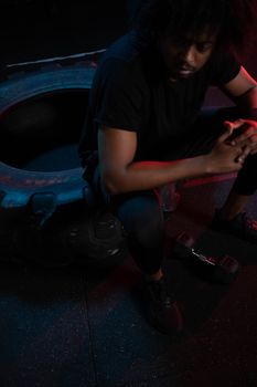 An athlete with afro hair rests on a tire in a dimly lit gym, clutching hands thoughtfully.