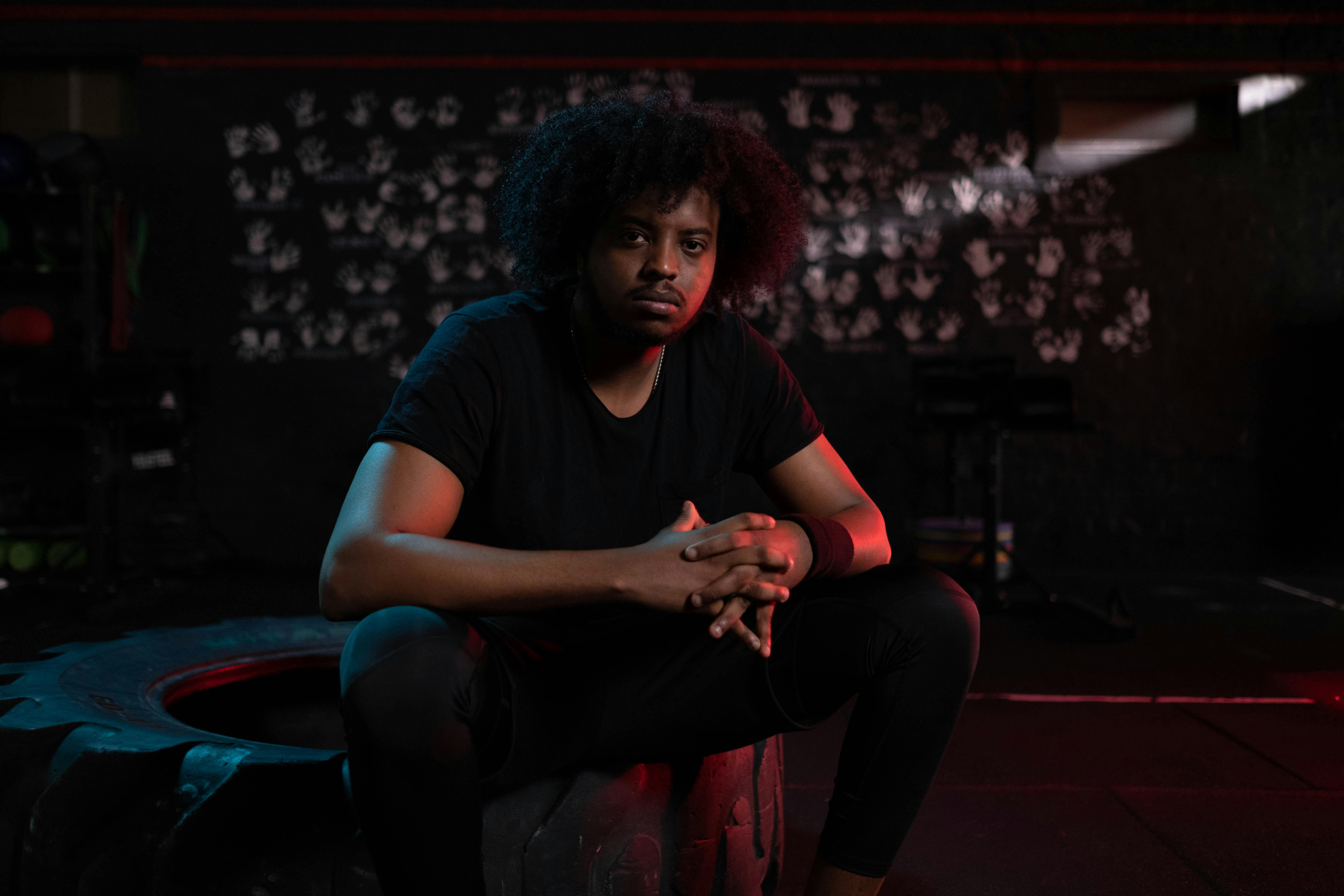 A thoughtful male athlete sits on a tire indoors with a moody gym ambiance.