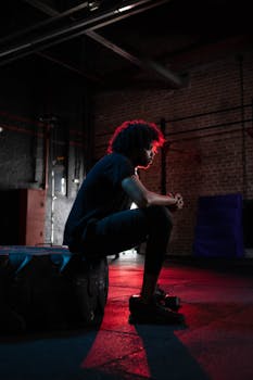 Side view of a man with afro hair sitting on a tire in a dimly lit gym with red lighting.