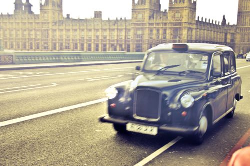 Free Classic London taxi speeding past the Houses of Parliament in a vibrant urban scene. Stock Photo