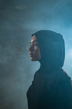 Side view of a contemplative woman in a hijab standing amidst a smoky background indoors.