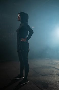 A Muslim woman in a hijab stretching indoors in a gym with a smoky background, highlighting fitness and wellness.