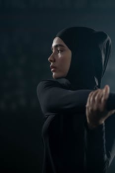 A focused Muslim woman in activewear stretches indoors, emphasizing fitness and wellbeing.