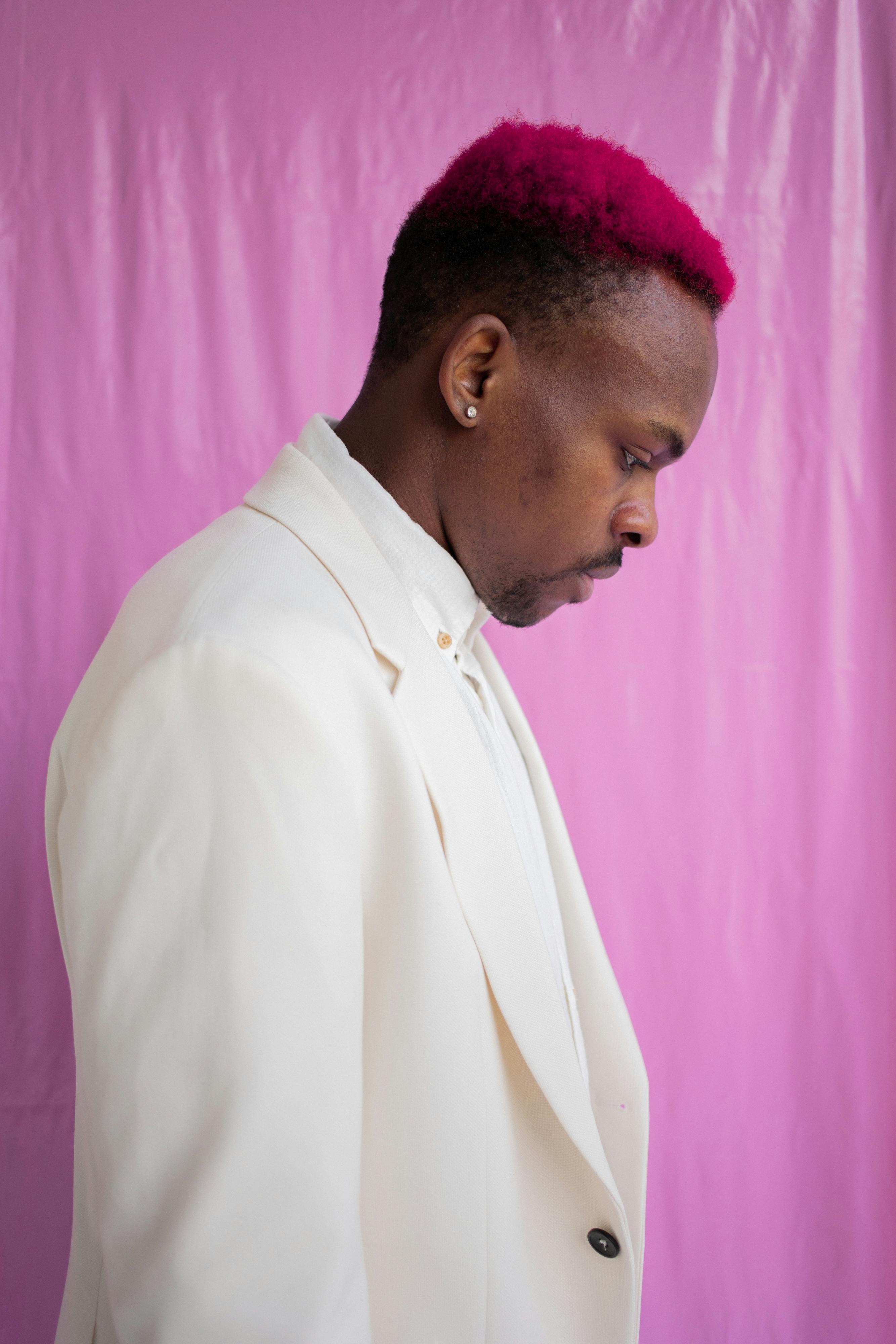 Side profile of a man in a white suit against a pink background, looking down.