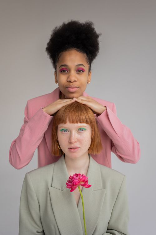 Free Multiracial female models with makeup in trendy clothes with pink flower in hand looking at camera on gray background in studio Stock Photo