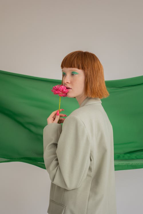 Side view of young female model with makeup in stylish outfit with pink flower in hand standing on gray background with green textile in studio