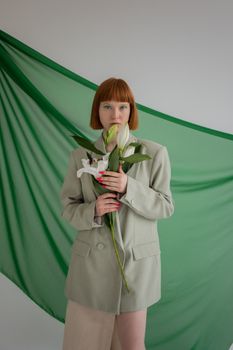 Attractive female in trendy clothes with flowers in hands looking at camera on gray background with green textile in studio