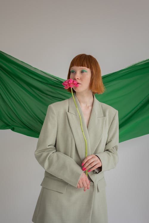 Gentle female with ginger hair and bright makeup wearing stylish blazer smelling blooming flower against green cloth hanging on gray background