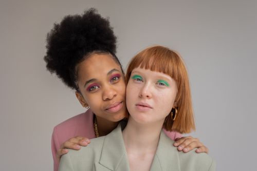 Free Gentle black woman with pink eyeshadows embracing red haired female from behind and looking at camera against gray background Stock Photo