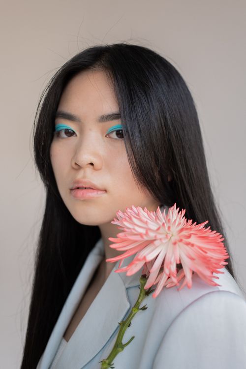 Free Charming Asian female with long dark hair in stylish blazer with blooming flower looking at camera against gray background Stock Photo