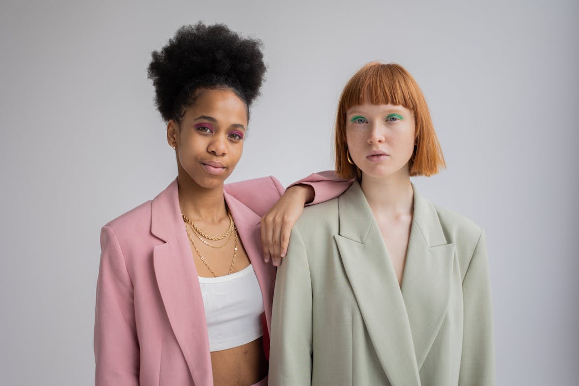 Stylish self assured African American female in trendy wear leaning on shoulder of red haired female with bright green eyeshadows on gray background