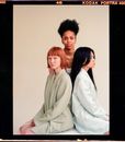 Emotionless diverse female models wearing stylish blazers sitting on cube against black woman on gray background
