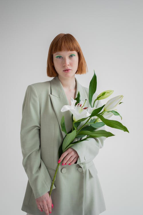 Free Confident red haired female with bright eyeshadows wearing blazer standing with blooming lily flowers and looking at camera against gray background Stock Photo