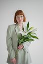 Confident red haired female with bright eyeshadows wearing blazer standing with blooming lily flowers and looking at camera against gray background