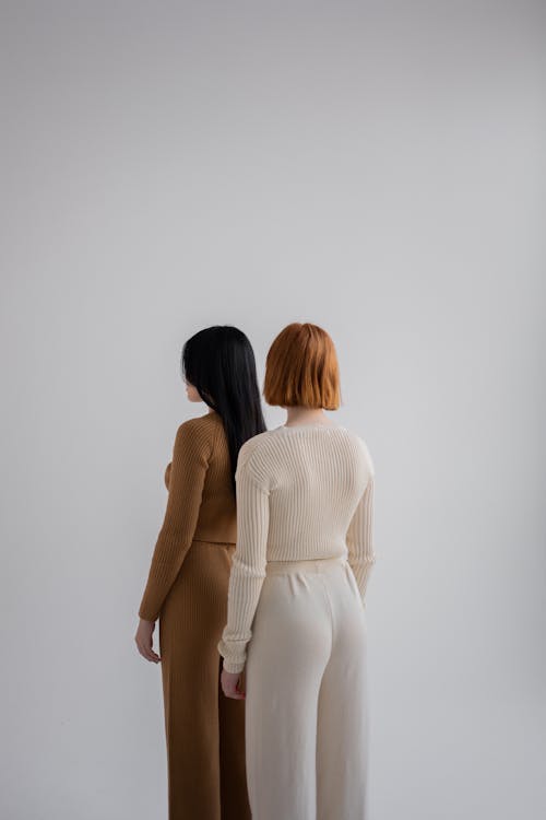Back view of unrecognizable female models wearing pants and sweaters standing together against gray background in studio