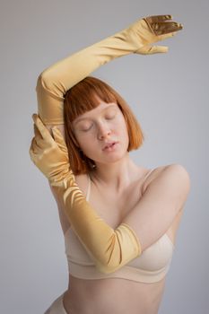 Young female with red hair closing eyes while gently touching raised hand in satin vintage gloves against gray background