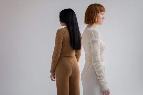 Back view of unrecognizable young female models wearing cozy simple suits while looking away and standing on white background in studio