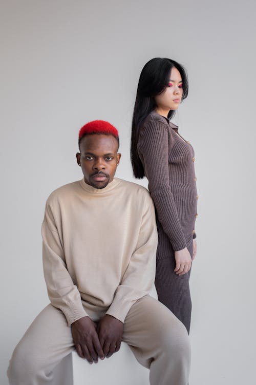 Confident African American male model in trendy outfit sitting near female standing on white background in studio