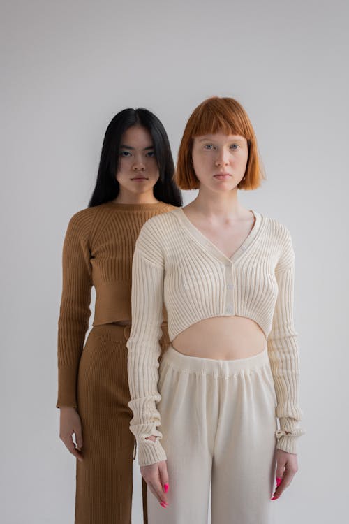 Emotionless female models in trendy clothes standing together while looking at camera on white background in studio