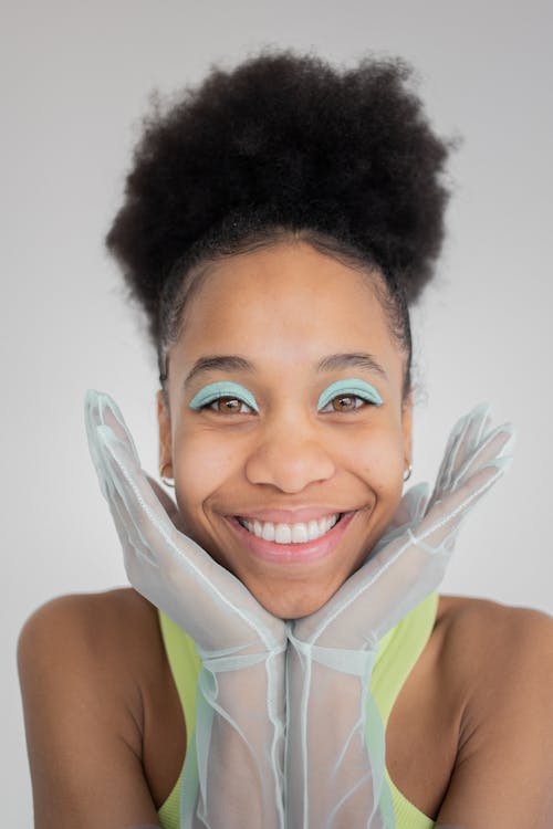 Cheerful smiling African American female with blue eyeshadows wearing transparent gloves and yellow top while putting face on hands and looking at camera