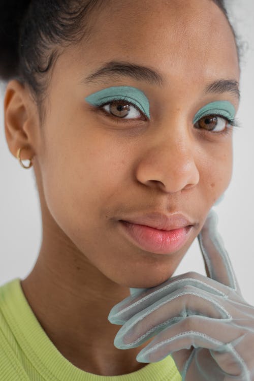 Young ethnic female with transparent gloves and with makeup of blue eyeshadows looking at camera and touching face with finger