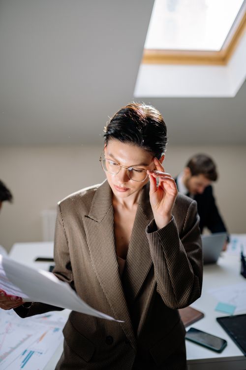 Free Focused woman in glasses reviewing paper documents in a sunlit office setting. Stock Photo